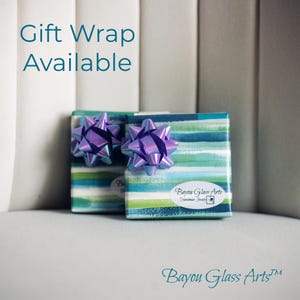 May include: Two small, square gift boxes wrapped in teal, blue, and green striped paper, each topped with a purple iridescent bow. The text "Gift Wrap Available" is in teal above the boxes. The boxes are labeled with "Bayou Glass Arts".