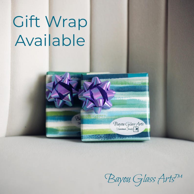 May include: Two small, square gift boxes wrapped in teal, blue, and green striped paper, each topped with a purple iridescent bow. The text "Gift Wrap Available" is in teal above the boxes. The boxes are labeled with "Bayou Glass Arts".