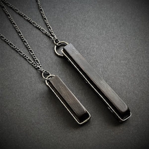 May include: Two silver-toned necklaces with rectangular, dark gray pendants. The pendants are encased in a silver frame and suspended from a delicate chain. The necklaces are displayed on a dark gray surface.
