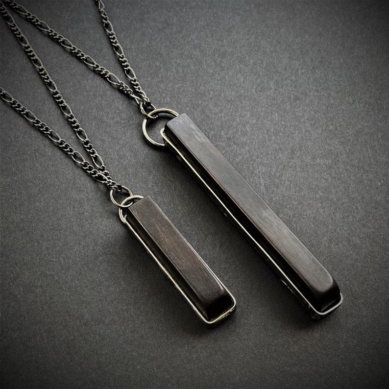 May include: Two silver-toned necklaces with rectangular, dark gray pendants. The pendants are encased in a silver frame and suspended from a delicate chain. The necklaces are displayed on a dark gray surface.