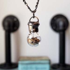 May include: A clear glass pendant necklace shaped like a lightbulb, filled with small gold-colored gears. The bulb is capped with a silver-colored metal and hangs from a twisted silver chain. The background is blurred, with black decorative elements.