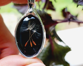 May include: Silver-toned pendant necklace with an oval-shaped, black resin center. The resin encases delicate white dandelion seeds with brown stems. The pendant hangs from a silver chain, creating a nature-inspired jewelry piece.