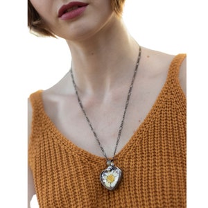 Puede incluir: Un collar de cadena plateada con un colgante en forma de coraz&oacute;n que contiene una margarita blanca prensada en su interior. El colgante est&aacute; en una persona que lleva una blusa de punto marr&oacute;n.