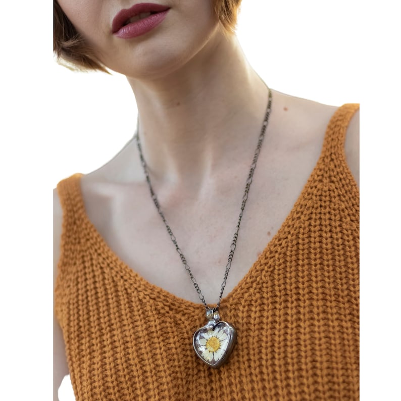 Puede incluir: Un collar de cadena plateada con un colgante en forma de coraz&oacute;n que contiene una margarita blanca prensada en su interior. El colgante est&aacute; en una persona que lleva una blusa de punto marr&oacute;n.