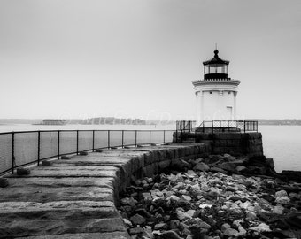 May include: Black and white photograph of a lighthouse on a stone pier. The white lighthouse has a black top and sits on a rocky foundation. A metal fence runs along the pier, and the ocean is in the background.