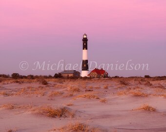 May include: A black and white lighthouse with a red-roofed building, set against a pink and purple sunset sky. The lighthouse is tall and cylindrical, with a light at the top. The foreground features sand dunes and dry grass.