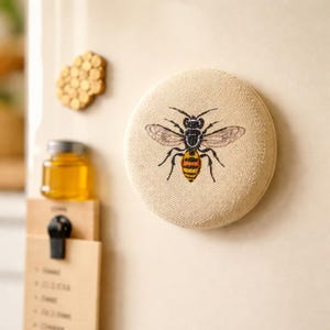 May include: A round, beige fabric magnet featuring an embroidered bee with black, yellow, and orange details. The bee has transparent wings. A wooden honeycomb and a jar of honey are also visible.
