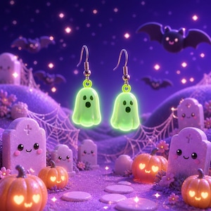 May include: A pair of dangling earrings featuring small, glowing light green ghost charms with black eyes and mouths. The charms are suspended from silver-toned metal fishhook wires, designed for pierced ears, showcasing a smooth, rounded finish.