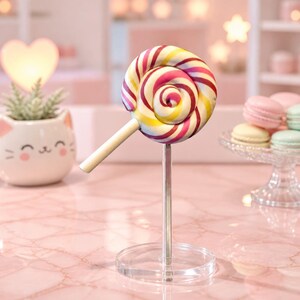 May include: A round, swirled lollipop featuring vibrant stripes of pink, yellow, and white, mounted on a clear acrylic stand. The sweet has a smooth, glossy finish and a white stick extending from the side, displayed as a decorative piece.