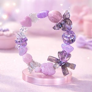 Pode incluir: Uma pulseira decorativa em forma circular, com uma variedade de contas roxas e transparentes. As contas incluem formas de coração, contas redondas e elementos cristalinos. A pulseira é exibida em um suporte rosa.