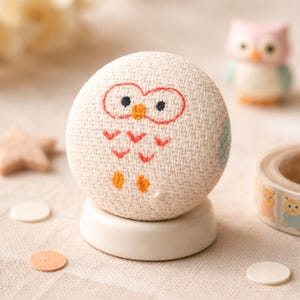 May include: A round, off-white owl-themed decorative object with embroidered details. The owl has red outlined eyes, a small orange beak, and orange feet. It sits on a white, circular base. Other owl-themed items are in the background.