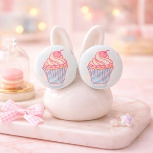 Puede incluir: Pendientes de cupcake azul claro con diseño de cereza, exhibidos en un soporte de cerámica blanco con forma de conejito. Los pendientes redondos presentan un diseño de cupcake con glaseado rosa y un forro azul. Fondo rosa suave.