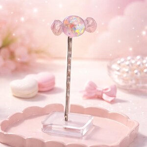 May include: A decorative candy-shaped object with a clear, iridescent centre and pink accents. The candy is mounted on a silver stem and a clear base. The background is a soft pink with other pink decorative elements.