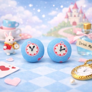 May include: Two blue fabric buttons with clock faces, featuring a white clock face with black hands and a pink border. The buttons are decorated with pink polka dots. The background includes a white rabbit figurine, a pocket watch, and a bottle labeled "Drink Me".