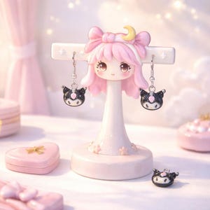 May include: A pink and white earring display stand shaped like a girl with pink hair and a bow. The stand holds two black and white cat-shaped earrings. A small pink heart-shaped box and a single earring are also visible.