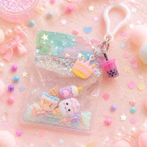 Op de afbeelding: Een doorzichtige plastic tas-sleutelhanger gevuld met iriserende glitters en kleine harsbedels, waaronder een lachend ijshoorntje en een marshmallow. De tas heeft een pastelkleurige bovenkant, een witte plastic clip en bedels van een kroon en boba-thee.