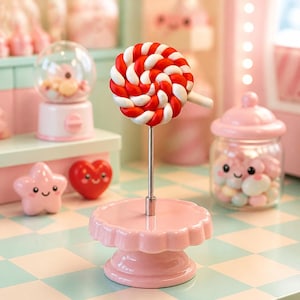 May include: A red and white swirled lollipop sits atop a silver metal rod attached to a round, scalloped-edge light pink pedestal stand. Nearby, a clear glass jar with a pink lid contains small sweets, alongside a pink star and a red heart.
