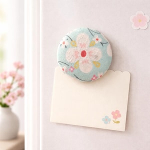 Puede incluir: Un imán redondo de tela con un diseño floral en azul claro, rosa y blanco, adherido a una tarjeta de notas crema en blanco. El imán tiene una flor central grande y pequeños detalles florales. También se ve una pequeña pegatina de flor rosa.