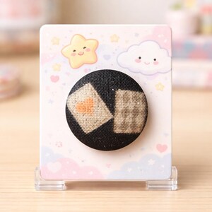 May include: A black fabric button with two embroidered squares, one with an orange heart. The button is attached to a decorative card with a pastel sky design, featuring a smiling star and cloud. The card is displayed in a clear plastic stand.
