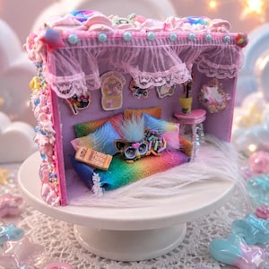 Peut inclure: Un diorama de pièce miniature présentant un canapé aux couleurs arc-en-ciel avec une petite créature à fourrure, un téléphone rétro et une table d'appoint. La structure est ornée de garnitures façon glaçage rose, de rideaux en dentelle et d'autocollants.