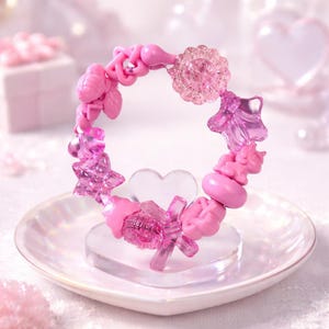 May include: A pink bracelet with various charms, including stars, flowers, and a unicorn. The bracelet is displayed on a heart-shaped stand and a white, heart-shaped dish. The charms are made of translucent and opaque pink materials.