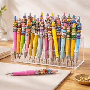 Pode incluir: Uma coleção de canetas coloridas com tampas de contas, exibidas em um organizador de acrílico transparente. As canetas têm barris de várias cores, incluindo rosa, amarelo e azul. Uma caneta rosa com uma tampa de contas repousa sobre uma superfície branca.