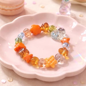 May include: A colorful beaded bracelet featuring orange star and flower-shaped beads, clear heart-shaped beads, and various other translucent beads in shades of orange, yellow, green, and blue. The bracelet is displayed on a pink, scalloped-edge plate.