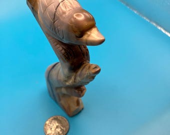 May include: A carved bird sculpture in shades of brown and tan perches on a branch. The bird has detailed feather markings and a curved beak. A coin rests near the base of the sculpture, providing a size reference. The sculpture is set against a blue background.