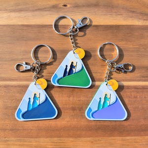 May include: Three triangular keychains with a religious scene. Each keychain features a silver keyring, a clasp, and a yellow accent. The artwork depicts figures in a landscape with blue, green, and purple backgrounds. The keychains are displayed on a wooden surface.