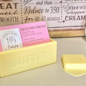 May include: A pale yellow rectangular butter dish with the word BUTTER embossed on the front. The dish holds a stack of pink business cards for Massey Makes Bakeshop. A small, separate pale yellow square piece resembling a pat of butter sits nearby.