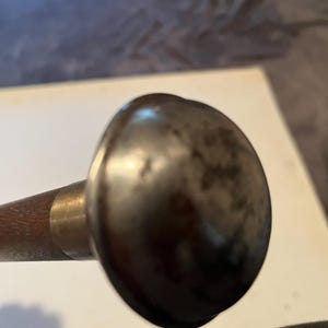 May include: A walking stick featuring a smooth, dark brown wooden shaft connected to a rounded, metallic handle. The handle has a polished, reflective surface with visible dark, mottled patina spots across its dome-shaped top.