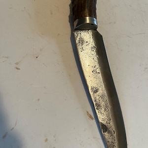 May include: A vintage hunting knife with a weathered, silver-colored blade and a dark brown, textured handle. The blade shows signs of wear and use. The handle appears to be made of antler material.