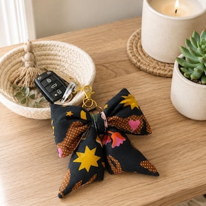 Peut inclure: Un porte-clés en forme de nœud papillon en tissu noir, avec une attache dorée, orné d'étoiles jaunes, de cœurs roses et de pois marron. Le porte-clés est sur une surface en bois, près d'un panier tressé et d'une bougie allumée.