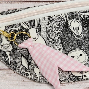 May include: A black and white zippered pouch with a whimsical animal print, featuring rabbits, owls, and other creatures. A pink gingham fabric bow is attached to a gold-colored keyring.