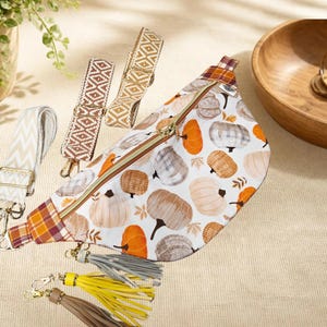May include: A white fanny pack featuring a pumpkin print in shades of orange, gray, and brown, with a gold zipper, plaid side accents, and three detachable tassels in gray, yellow, and brown, plus two interchangeable patterned shoulder straps.