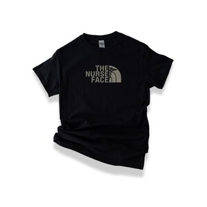 May include: Black t-shirt featuring the text "THE NURSE FACE" in grey, with a caduceus symbol incorporated into the design. The shirt has a crew neck.