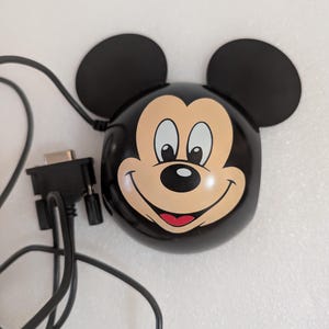 May include: A black Mickey Mouse-shaped device with attached black ears and a smiling face. A black cable and connector extend from the device. The device is on a white surface.