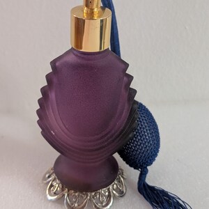 May include: A frosted deep purple glass perfume bottle with a scalloped, ribbed body and a gold-toned metal collar. The bottle features a blue fabric-covered atomizer bulb with a matching blue tassel and a decorative silver-toned metal base.
