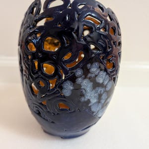 May include: A dark blue ceramic vase with intricate cut-out patterns. The vase has a glossy finish and a textured surface with a lighter interior. The design includes floral and abstract shapes, creating a decorative and artistic piece.