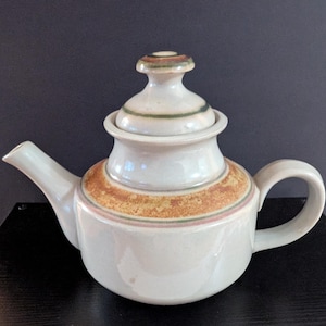 May include: A ceramic teapot with a light cream-colored glaze, featuring a wide band of mottled brown and orange texture around the shoulder. The lid has a matching knob with thin green and brown rings, and the spout and handle are curved.
