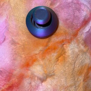 May include: A small, round, iridescent bottle stopper with a curved handle. The stopper has a metallic sheen, displaying shades of blue, purple, and green. The background is a soft, plush fabric in a blend of pink, orange, and yellow.