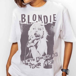 May include: White t-shirt featuring a black and white graphic of a singer with the word "BLONDIE" above and "NORTH AMERICAN TOUR 1982" below. The shirt is worn by a person wearing black shorts.