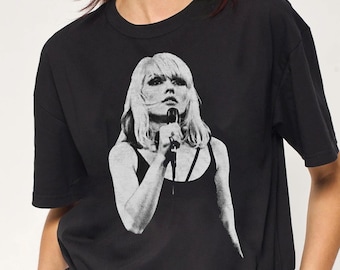 May include: A black short-sleeved t-shirt featuring a white graphic print of a person with shoulder-length hair holding a microphone. The graphic depicts a person wearing a tank top, captured in a monochromatic, high-contrast photographic style.