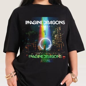 Puede incluir: Camiseta negra con la banda Imagine Dragons. La camiseta muestra el nombre de la banda en blanco, junto con las palabras "Origins" y "Evolve". Un gráfico colorido de una persona en un círculo con un rayo de luz arcoíris está en el centro.