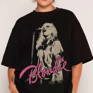 Könnte beinhalten: Schwarzes T-Shirt mit einem Graustufenbild einer Sängerin und eines Mikrofons sowie dem pinkfarbenen Wort "Blondie". Das T-Shirt hat einen klassischen Rundhalsausschnitt.