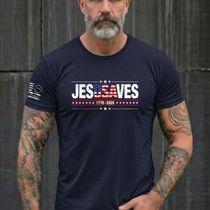 May include: A navy blue short-sleeved t-shirt featuring the white text JESUSA VES with the letters USA stylized as an American flag. Below the text is 1776-2026 in white, flanked by red and white stripes and blue stars. A small flag emblem is on the sleeve.