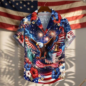 Puede incluir: Una camisa de manga corta con botones y un estampado patriótico vibrante que muestra un águila calva, la Estatua de la Libertad, la Campana de la Libertad, fuegos artificiales, rayos y flores de hibisco rojas, blancas y azules sobre un cielo estrellado.