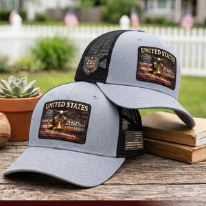May include: Two gray baseball caps with black mesh backs. Each cap features a rectangular patch displaying an American flag, a bell, and the text "UNITED STATES 250TH ANNIVERSARY". One cap also has a small shield-shaped patch reading "250 YEARS".