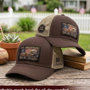 May include: Two brown baseball caps with tan mesh backs. Each cap features a rectangular patch displaying an American flag, a bald eagle, and the text 250TH ANNIVERSARY We the People. One cap has a circular 1776-2026 patch on the side mesh.