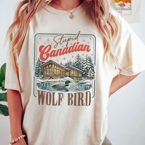 May include: A cream-coloured t-shirt with a graphic design. The design includes the text "Stupid Canadian" above a lakeside cabin illustration and a loon. The words "WOLF BIRD" are below. The design has a vintage, outdoorsy aesthetic.